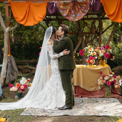 Bride in a lace veil and groom in a green velvet suit kiss under an orange and floral draped arbor