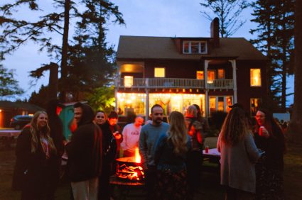 A group gathers around a fire pit at dusk in front of the lodge's lit windows