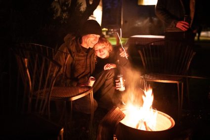 A couple sits close together by a fire pit at night roasting a marshmallow