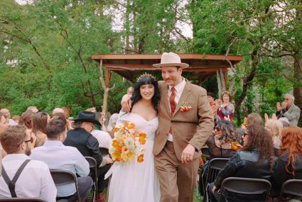 Bride and groom walk back down the aisle past cheering seated guests under a wooden arbor