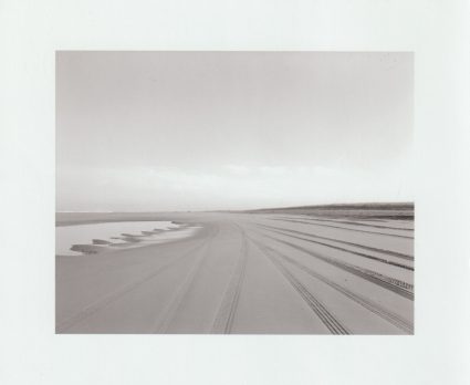 Black and white photograph of tire tracks and tide pools stretching across a flat beach toward the horizon