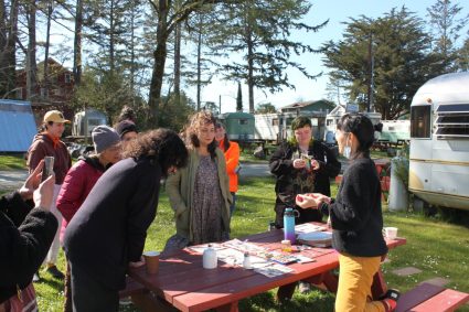 A group gathers around a picnic table outdoors near vintage trailers on a sunny day