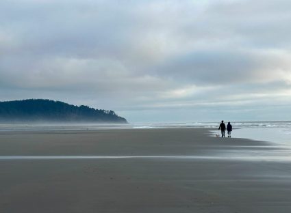 Two silhouetted figures walking along a foggy beach with a headland in the distance
