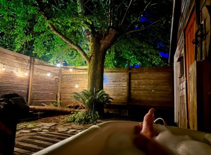 Outdoor soaking tub at night with a foot resting on the rim, string lights overhead