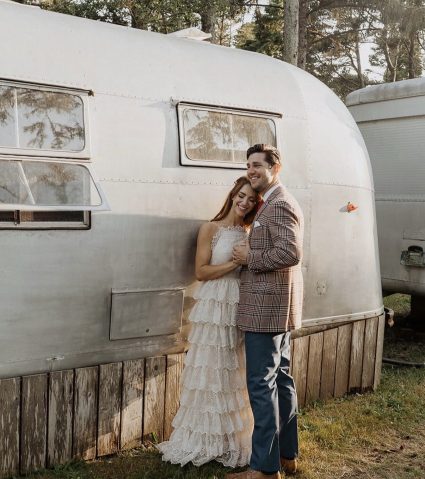 Bride and groom lean against a vintage silver Airstream trailer at sunset