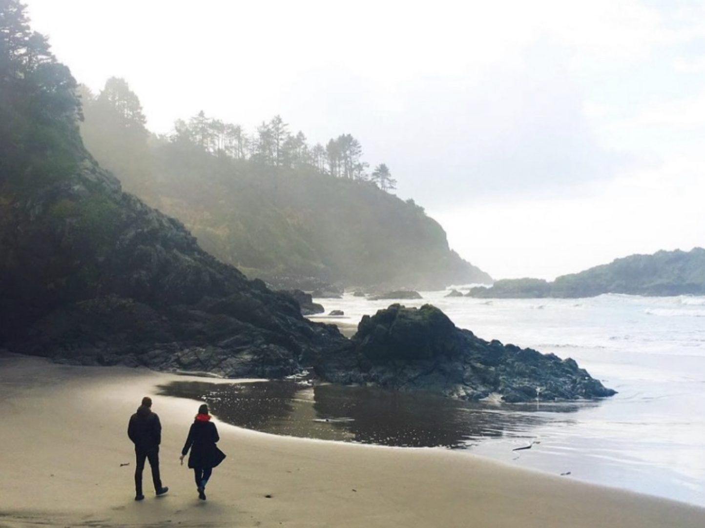 Two people walk along a foggy beach past dark rock formations at the base of a forested bluff