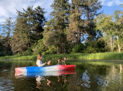 A child paddles a red white and blue kayak with a small dog standing at the bow on a calm pond