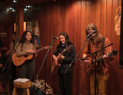 Three musicians in a wood paneled room singing together at microphones with acoustic guitars