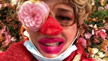 A woman's face close up with a pink flower covering one eye, red paint on her nose, and a surgical mask, lying in grass