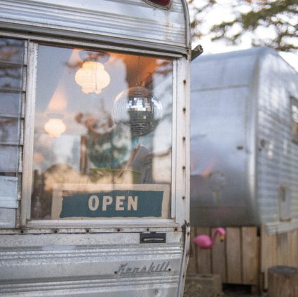 Window of a vintage trailer reflecting string lights and a disco ball with an OPEN sign propped inside