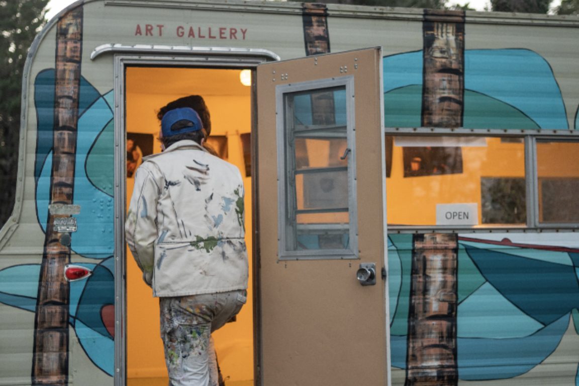 Vintage trailer painted with a teal mural and Art Gallery sign, an OPEN sign in the window as a visitor steps inside