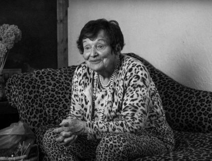 An older woman with short hair smiles while sitting on a leopard print couch wearing a matching leopard print outfit