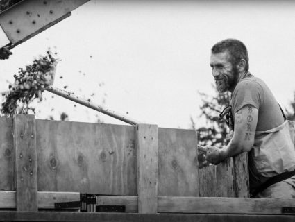 A bearded man in an apron builds a wooden crate outdoors as wood shavings fly