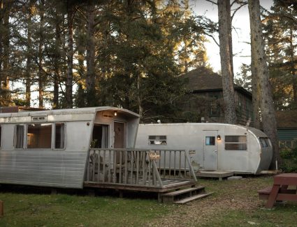 Two vintage aluminum travel trailers joined by a wooden deck, parked among tall pines at dusk