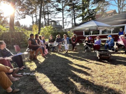 A group sits in a wide circle of chairs outdoors near a vintage trailer at sunset
