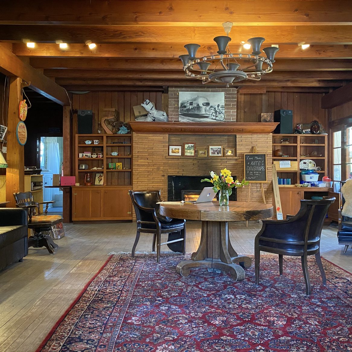 Lodge living room with exposed wood beams, a stone fireplace, wagon-wheel chandelier and a red patterned rug