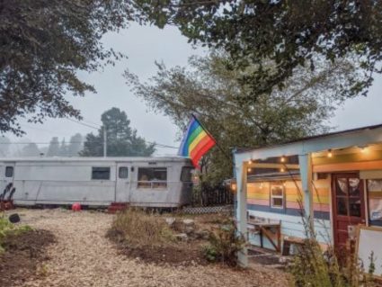 A vintage silver trailer sits beside a striped cabin decorated with string lights and a rainbow flag