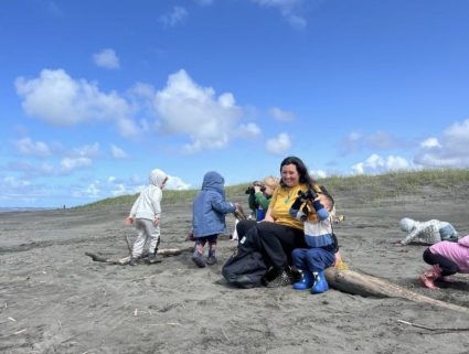 A woman sits with young children on a sandy beach near dune grass under a partly cloudy sky