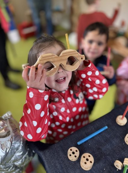 A girl playfully holds wooden toy glasses up to her eyes at a craft table with wooden pieces