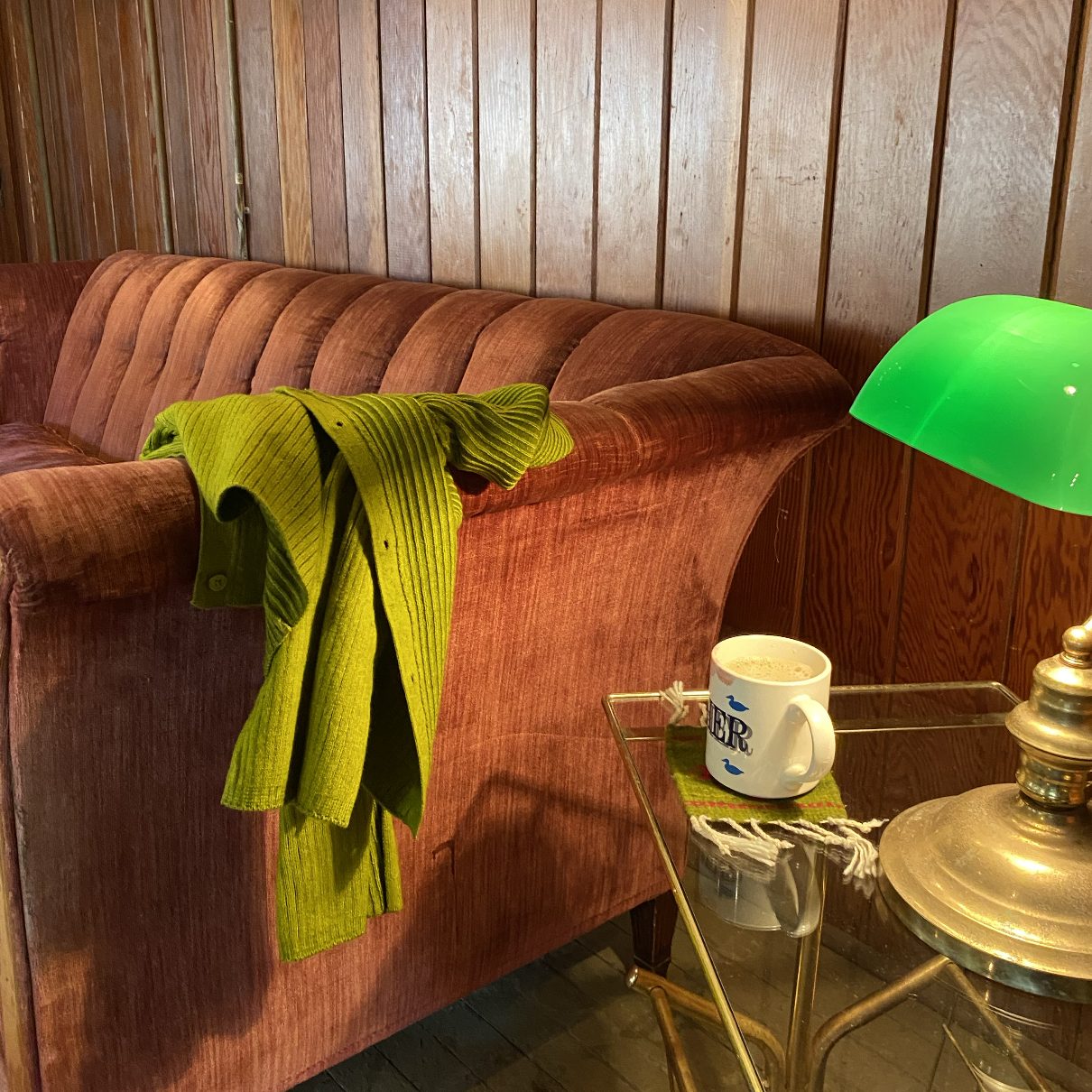 A green cardigan draped over a rose velvet channel back sofa beside a brass lamp and glass bar cart
