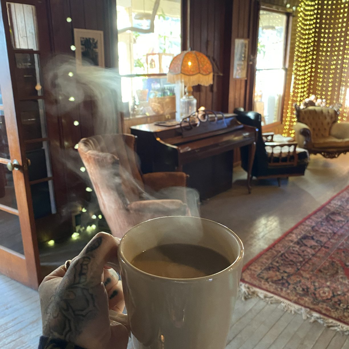 A steaming ceramic mug held in a tattooed hand with a cozy wood paneled living room blurred behind it