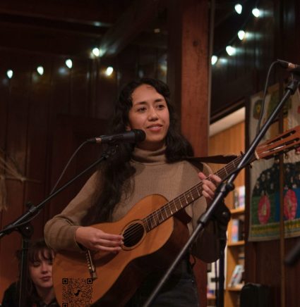 Woman singing on stage with an acoustic guitar under string lights inside the lodge