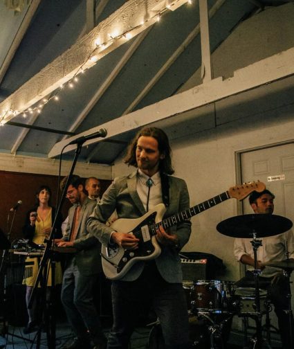 Band performing under string lights indoors, with guitarist, singer, and drummer on stage