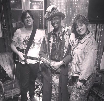 Black and white photo of three musicians posing together after a show, one wearing a feathered hat