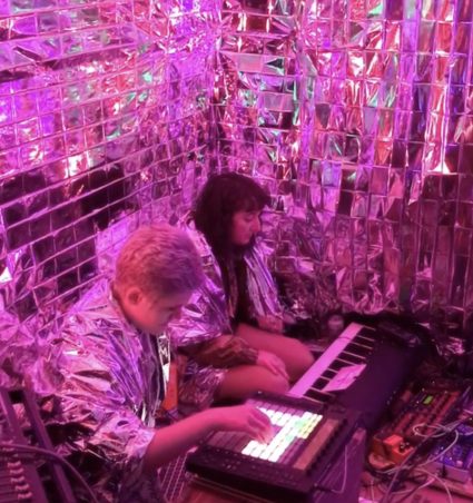 Two musicians sit inside a small room covered in shredded silver mylar, playing a keyboard and sampler under pink light