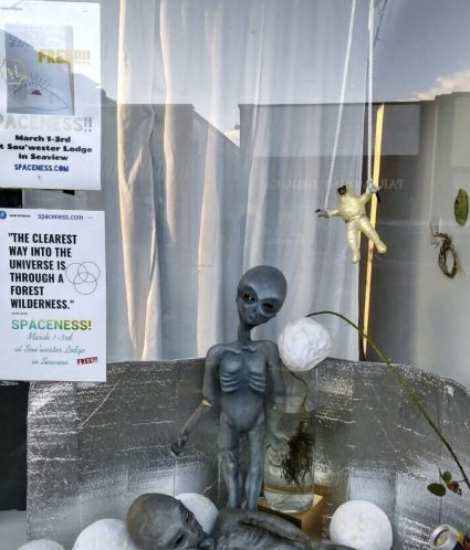 A shop window display of grey alien figurines, foil balloons, and a flyer advertising the Spaceness free art event at Sou'Wester Lodge
