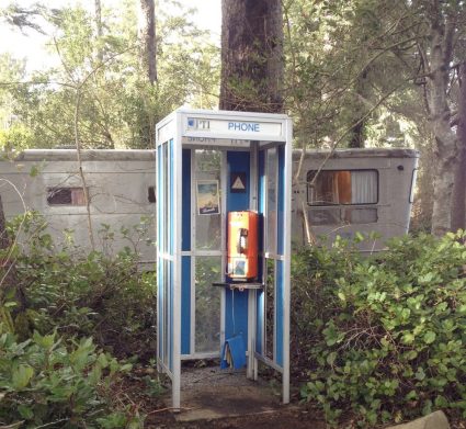 A vintage blue telephone booth marked PHONE standing in overgrown brush beside a trailer