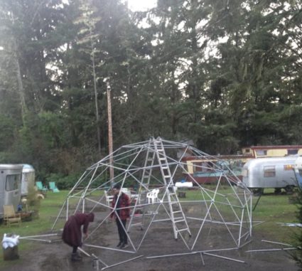Two people building a large geodesic dome frame from metal rods in a grassy campground among vintage trailers