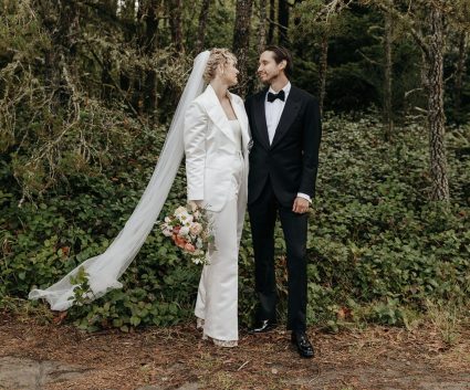 Bride in a white suit and veil walks beside the groom in a black tuxedo through the forest
