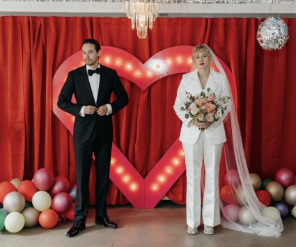 Bride in a white suit and groom in a tuxedo stand in front of a lit red heart backdrop with balloons