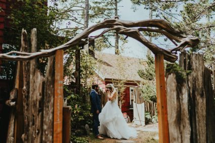 Bride and groom framed under a rustic wood and branch arbor with the red lodge building behind them
