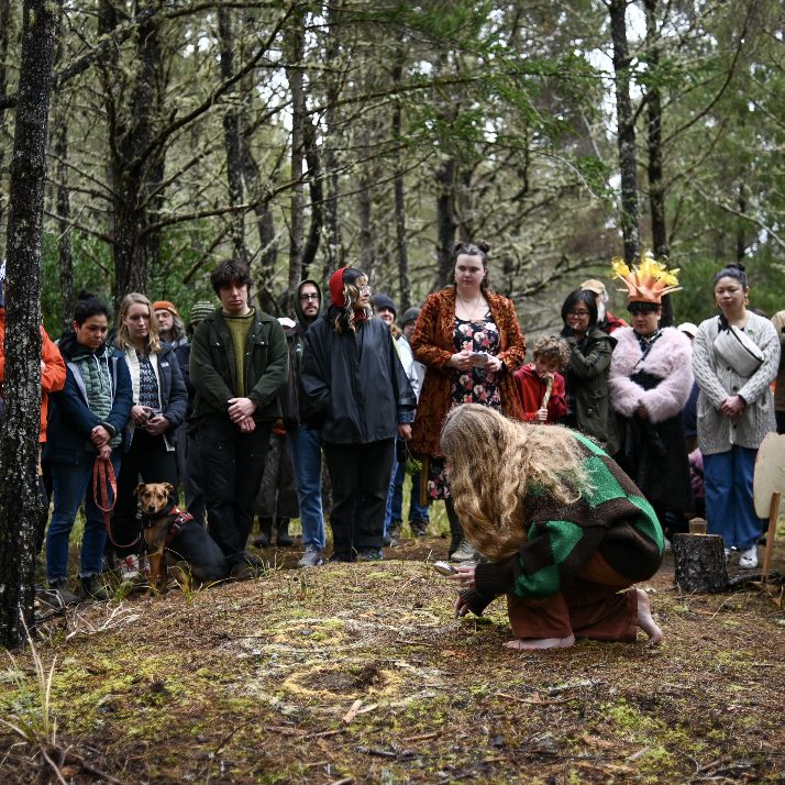 A crowd in the forest watches a performer in a green sweater kneel on the ground during a ritual-like scene