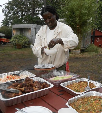 A woman in white serves food from foil catering trays on an outdoor table