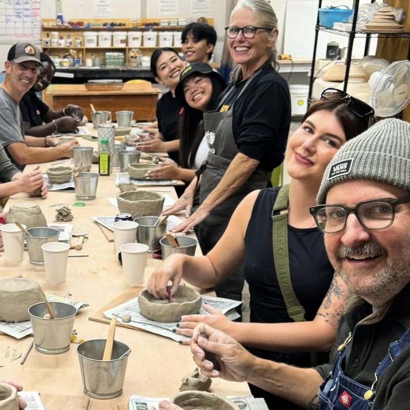 A group of adults smile at a long table during a pottery class, shaping clay bowls and mugs