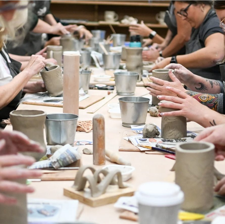 Hands shaping clay cups and small sculptures at a long communal pottery table