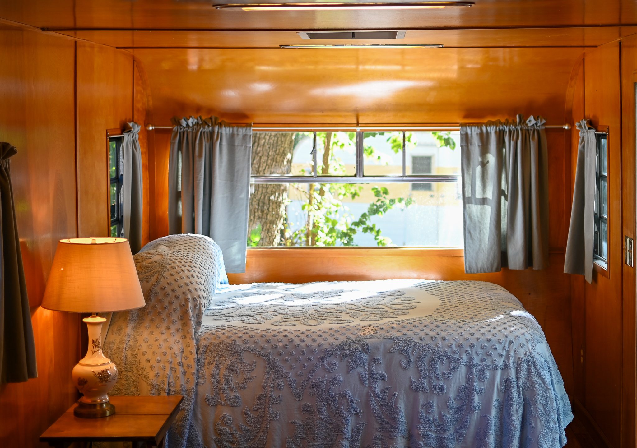 Vintage travel trailer bedroom with polished wood walls, gray curtains, and a bed by a large window