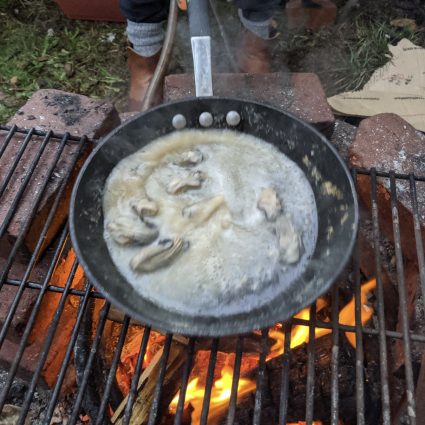 A cast iron pan of oysters cooks over an open charcoal fire on an outdoor grill