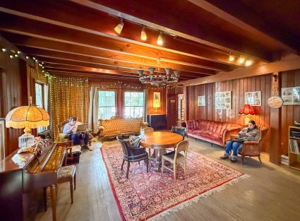 A wood beamed lodge living room with string lights, a piano, red velvet sofas and a round table on a rug