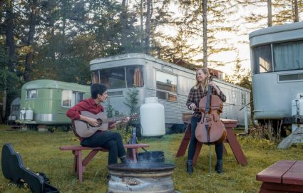 A man plays guitar and a woman plays cello outdoors among vintage trailers at dusk