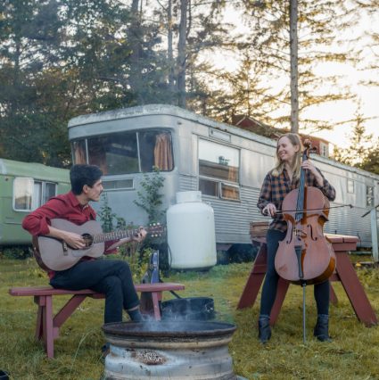 Musicians playing guitar and cello beside a fire pit in front of a vintage trailer among trees