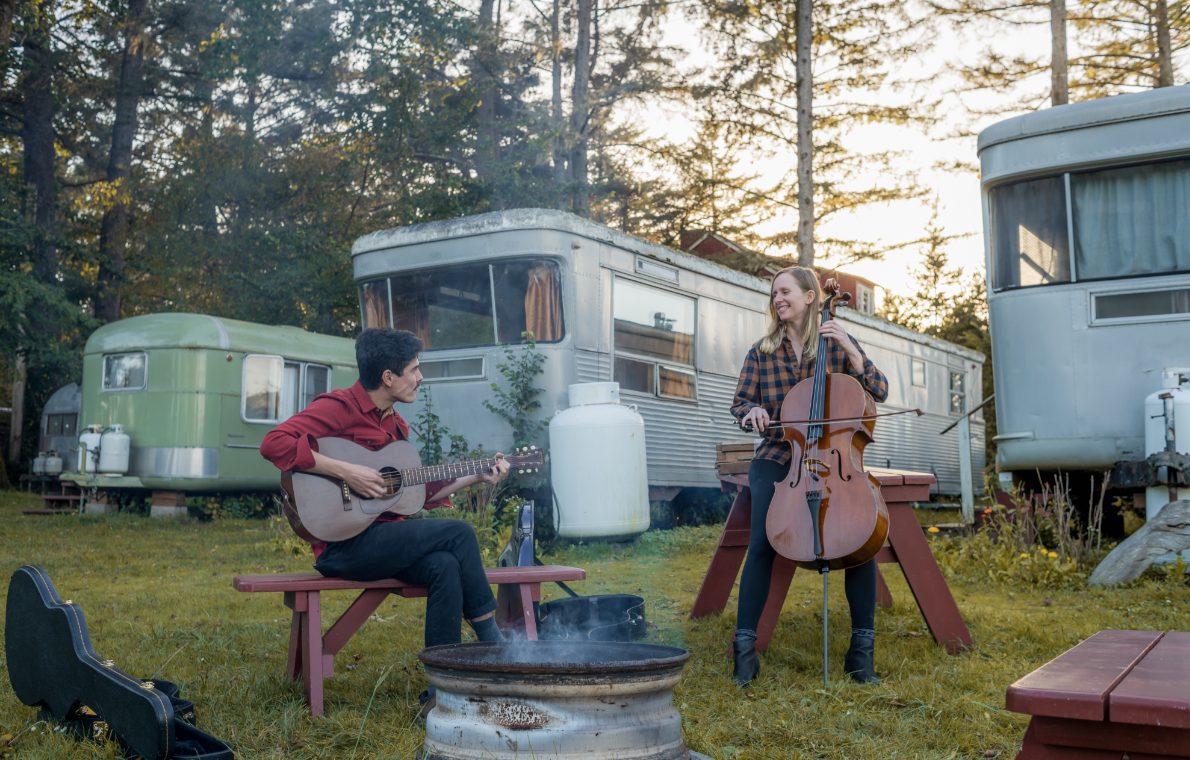 Man playing guitar and woman playing cello together by a fire pit among trailers