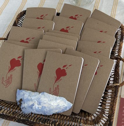 A woven basket filled with kraft paper notebooks stamped with a red flower and hands design